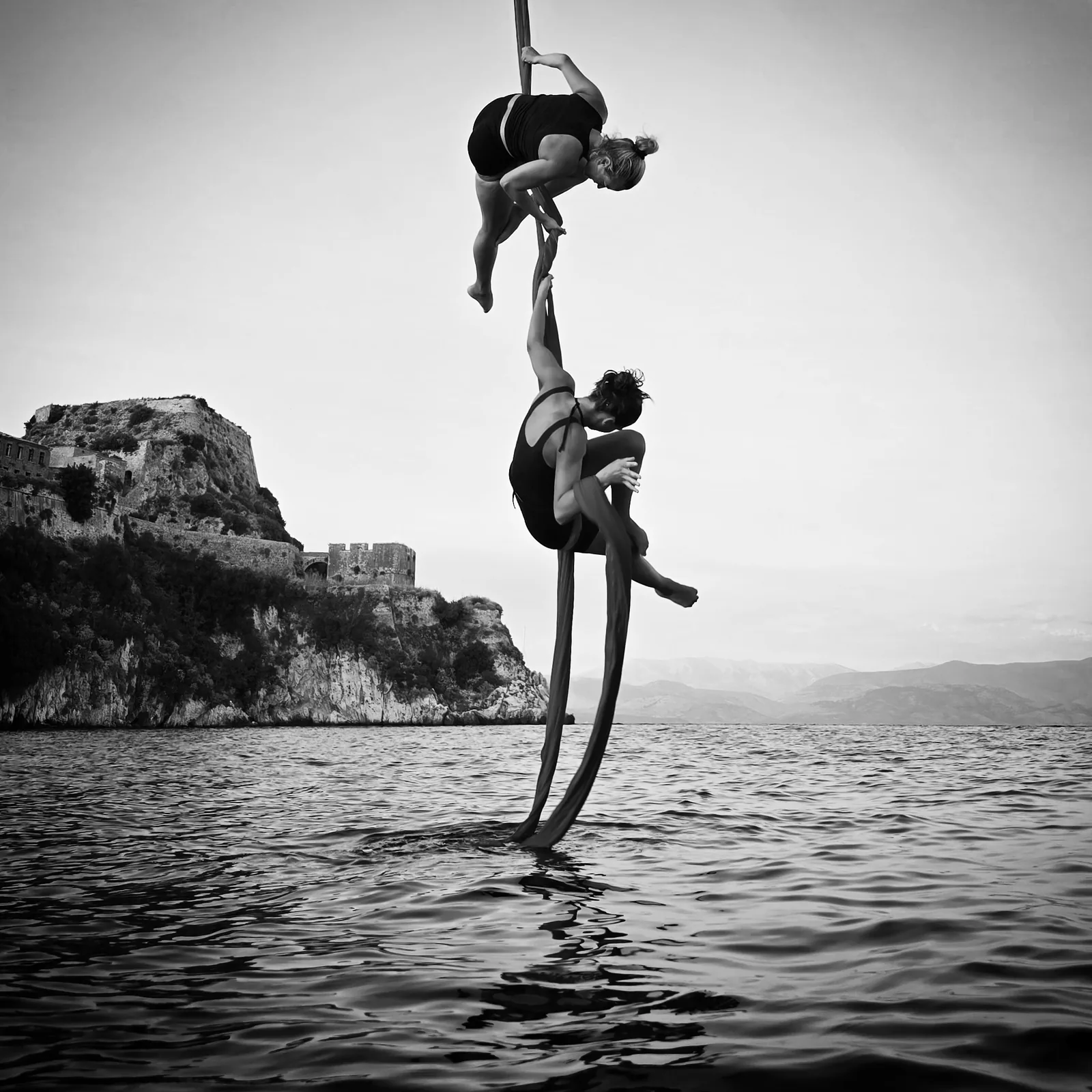 Two aerialists on silks above the water in black and white, a cliff fortress behind