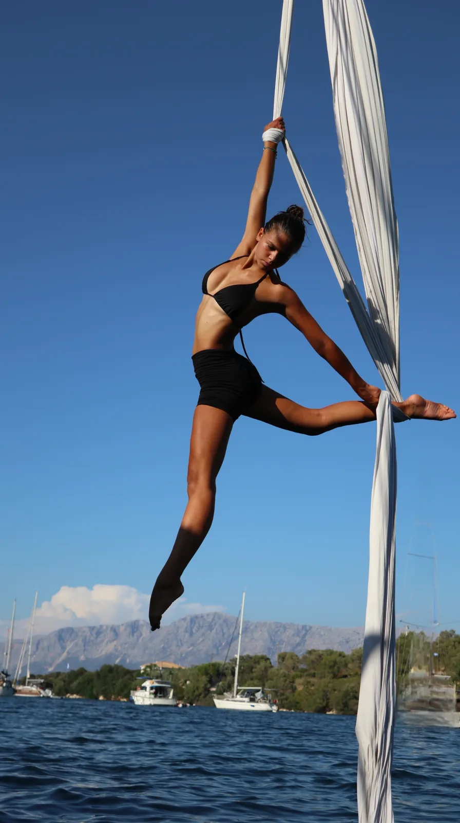 Standing split on white silks above a bay, sailing boats in the distance